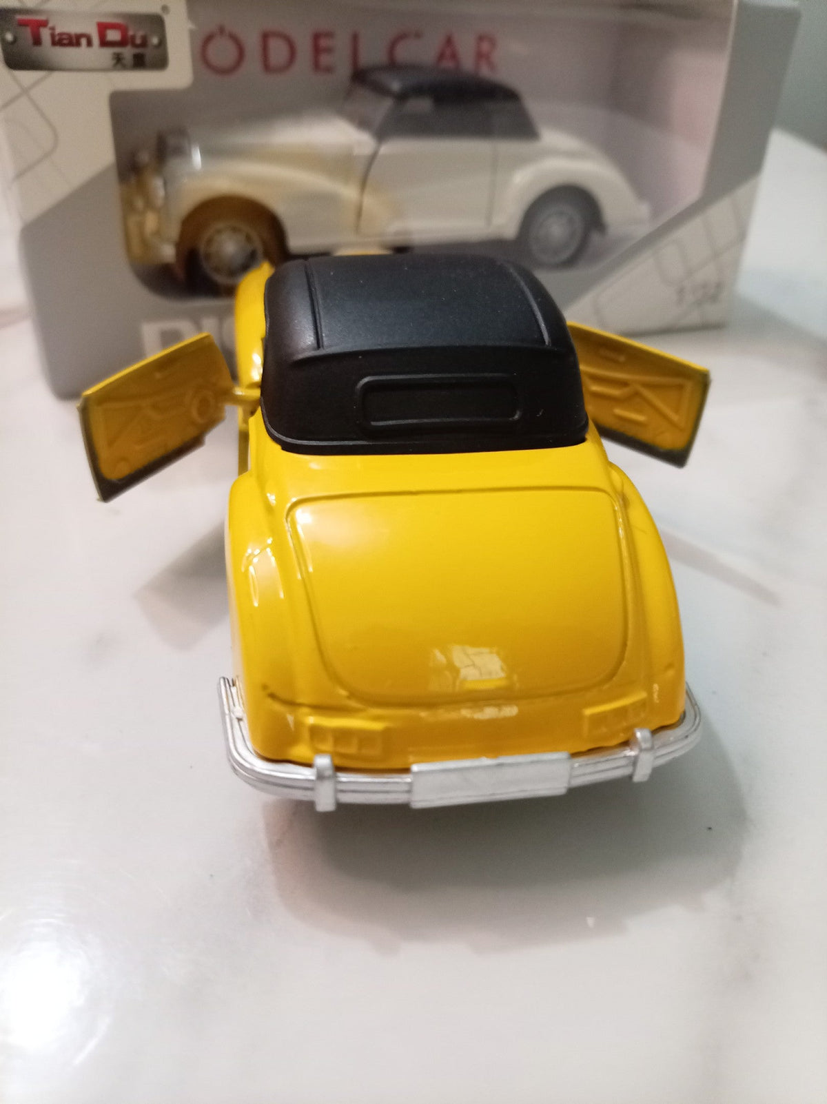 A yellow vintage model car with a black roof and open doors, shown from the rear, with its retail packaging box in the background.