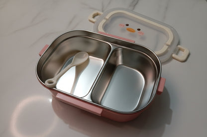 A stainless steel two-compartment lunch box in pink color body, shown open on a light marble-like surface. A white plastic spoon rests inside the left compartment, and a decorative lid with a cartoon face is partially visible behind it.