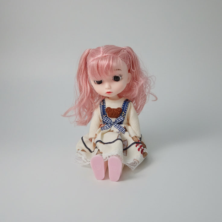 A doll with long pink hair sits on top of a black and yellow box on a tabletop. The doll wears a cream dress with blue gingham straps, a brown teddy bear motif, and lace trim.
