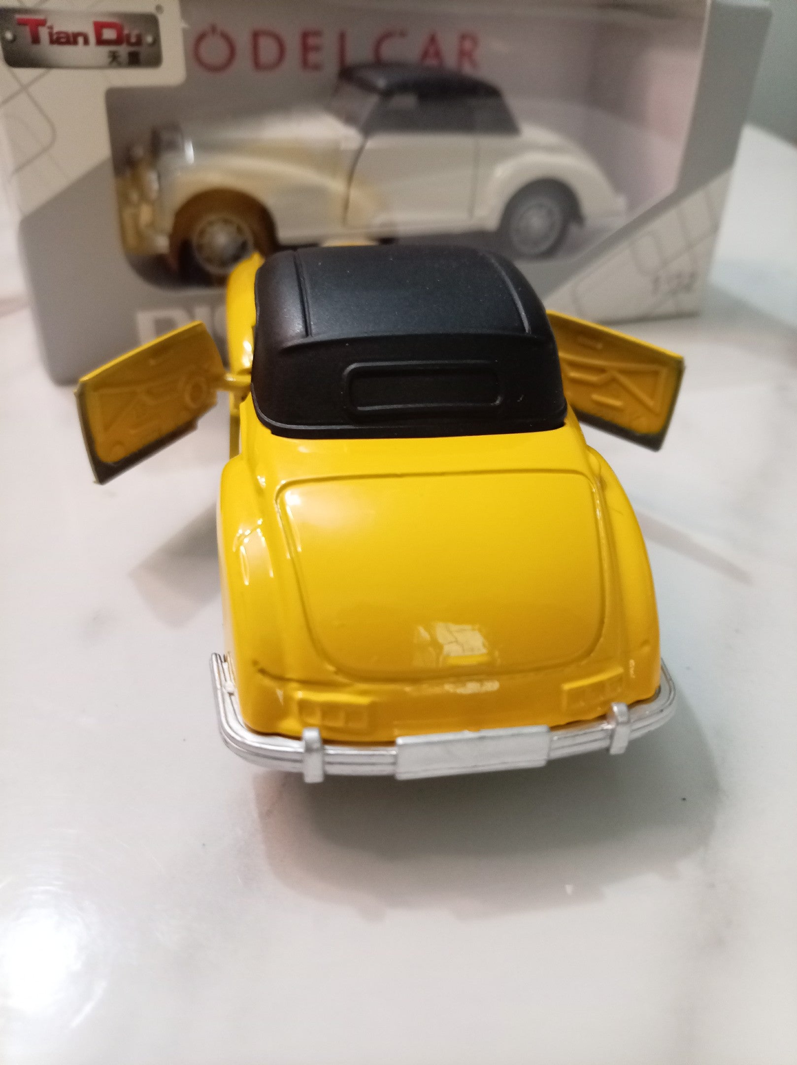 A yellow vintage model car with a black roof and open doors, shown from the rear, with its retail packaging box in the background.