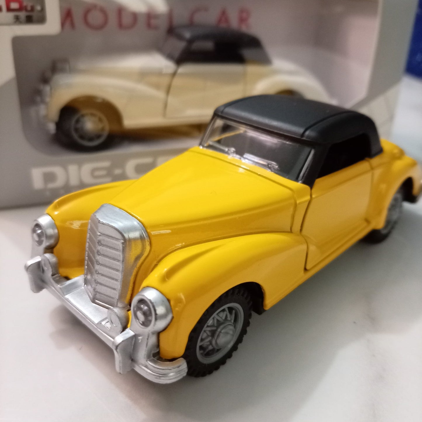 A yellow vintage model car with a black convertible top and silver chrome grille, shown in front of its white and gray packaging box.